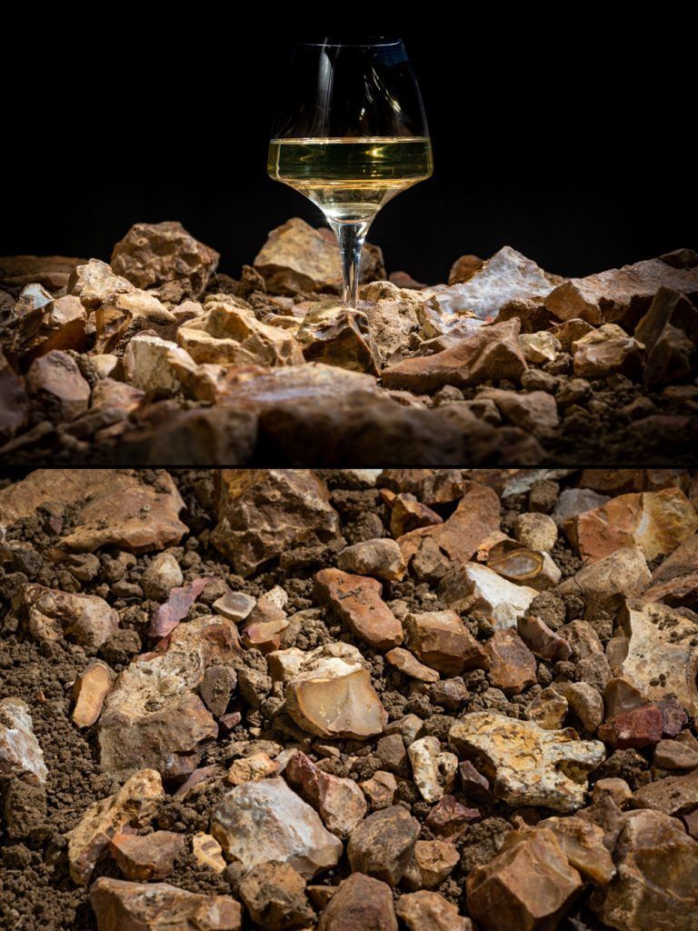 Wine glass filled with white liquid, set amidst various-sized rocks and dirt on a dark background.