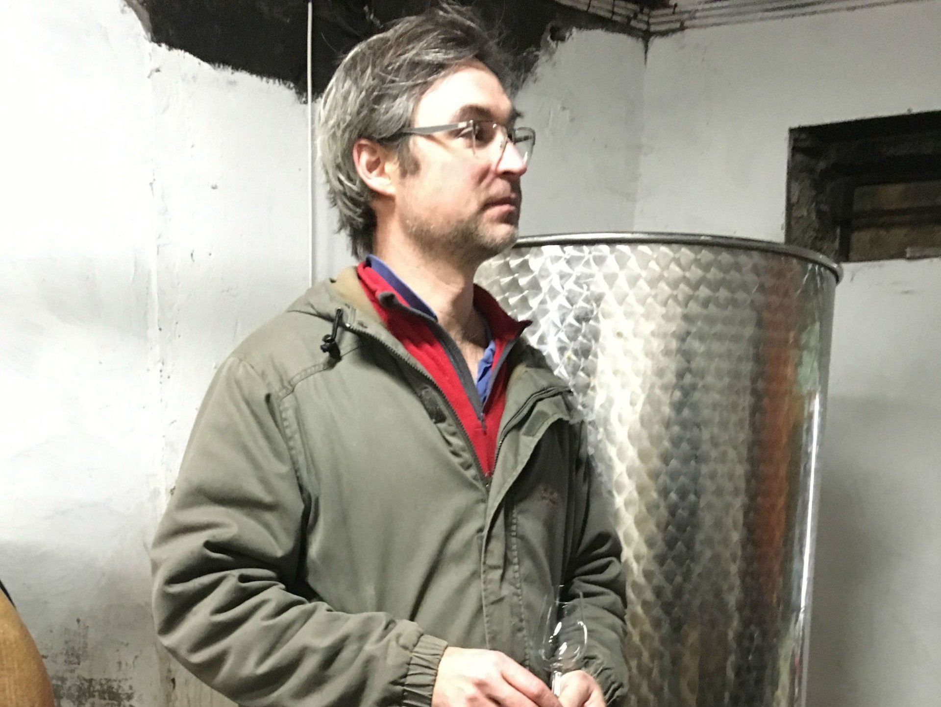 Man in glasses and jacket stands near a large metal tank, at Domaine Les Cortis