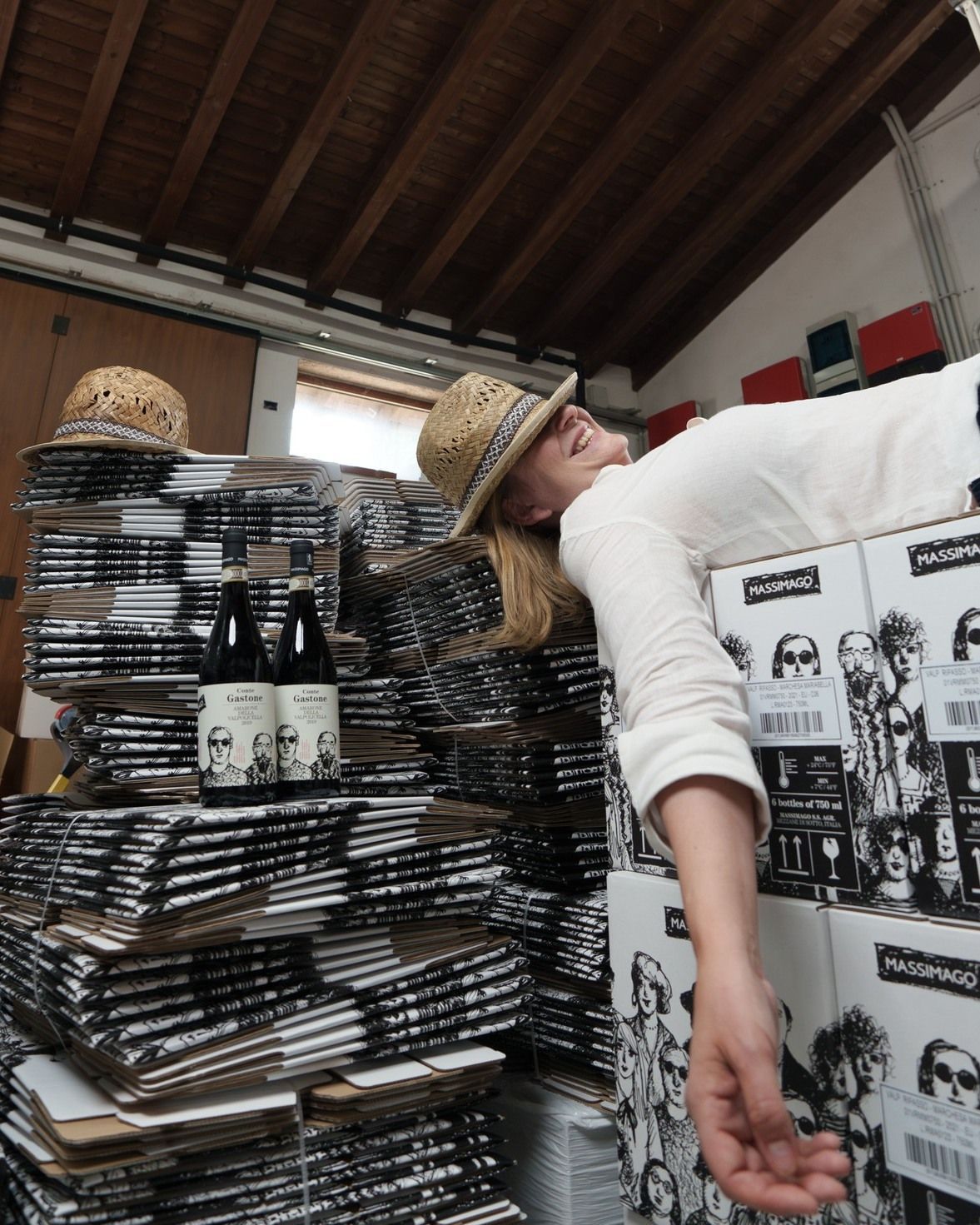 Massimago - A woman (Camila) in straw hat rests on boxes, two wine bottles on cardboard stacks in a warehouse.