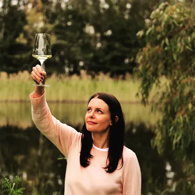 Woman holding a glass of Di Giovanna white wine up to the light, lake and trees in background.