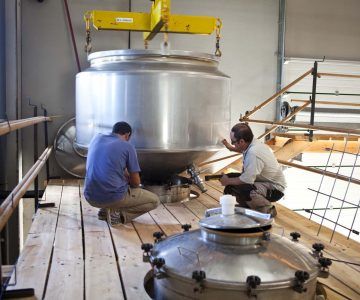 Two workers installing a large silver tank, suspended by a crane, on a wooden platform, at Pago Casa Gran.
