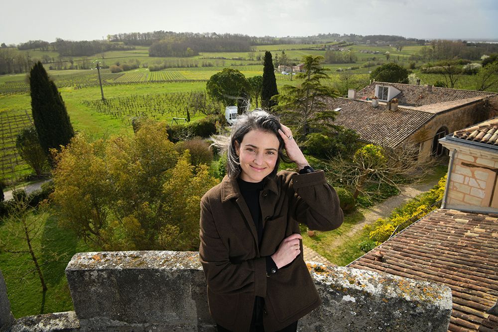 Peybonhomme-les-Tours - Woman on a stone wall smiles, hand in hair, overlooking green landscape with vineyards and buildings.