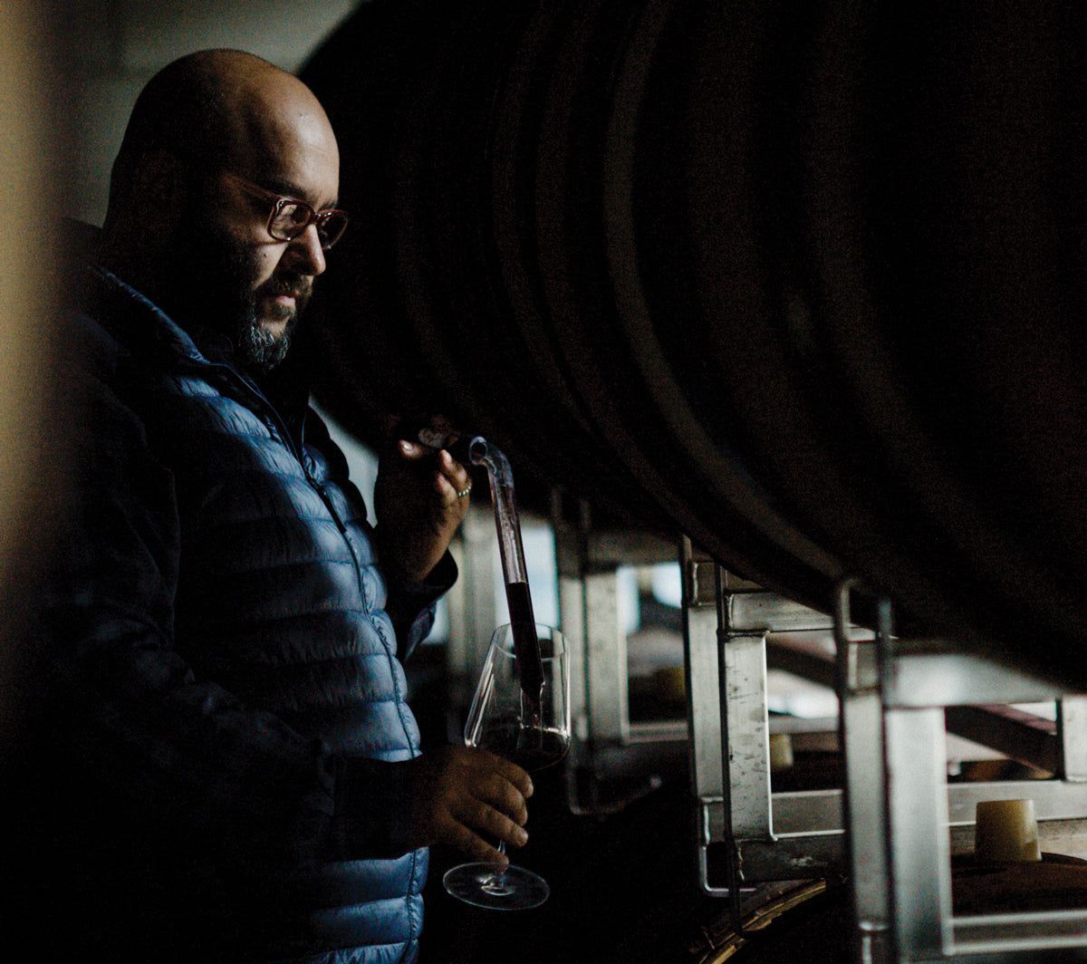 Man in Sandhi cellar, tasting wine from a barrel with a glass pipette.