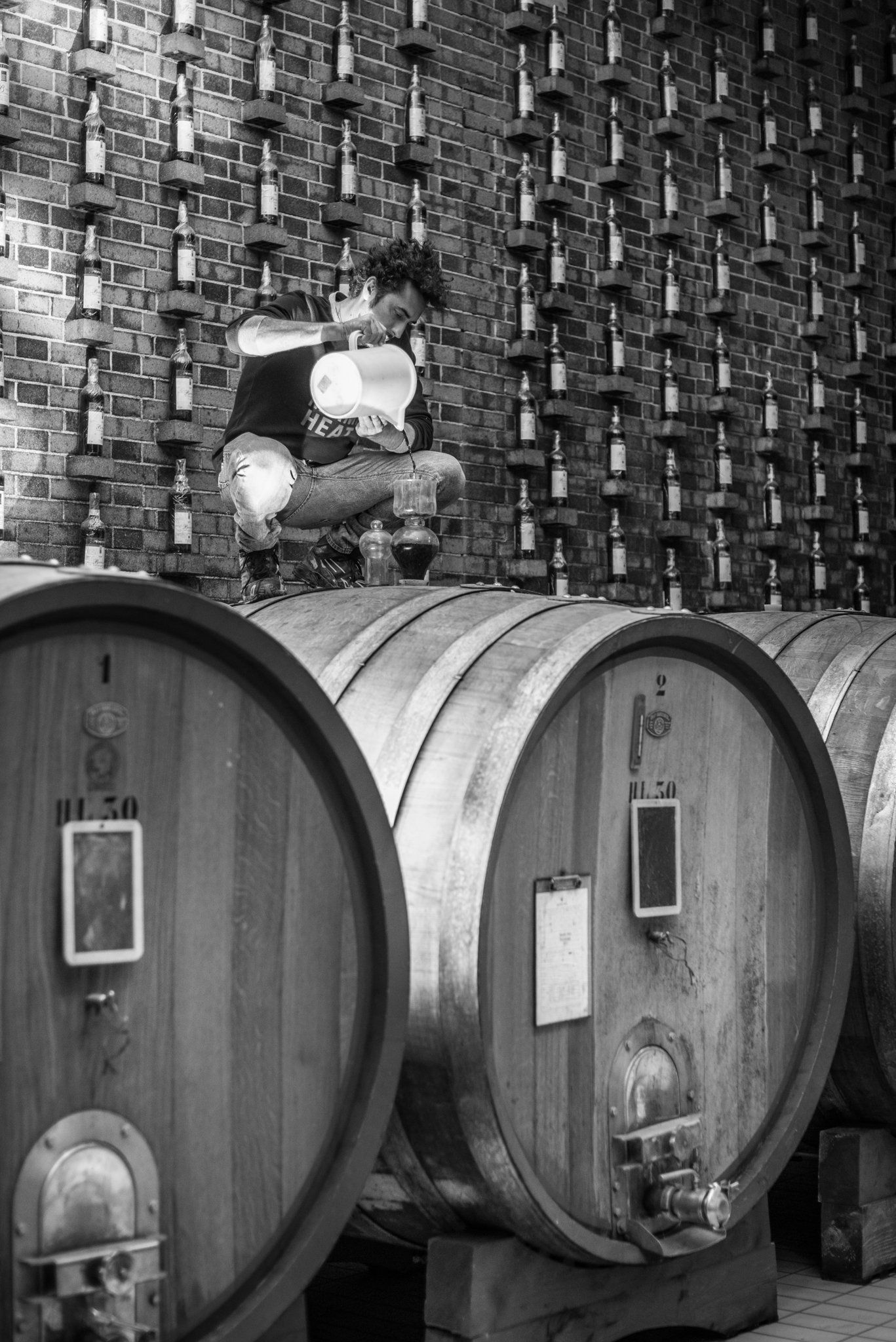 Man inspecting wine, squatting atop barrels in a cellar with wine bottle displays on a brick wall, at Rivetto.