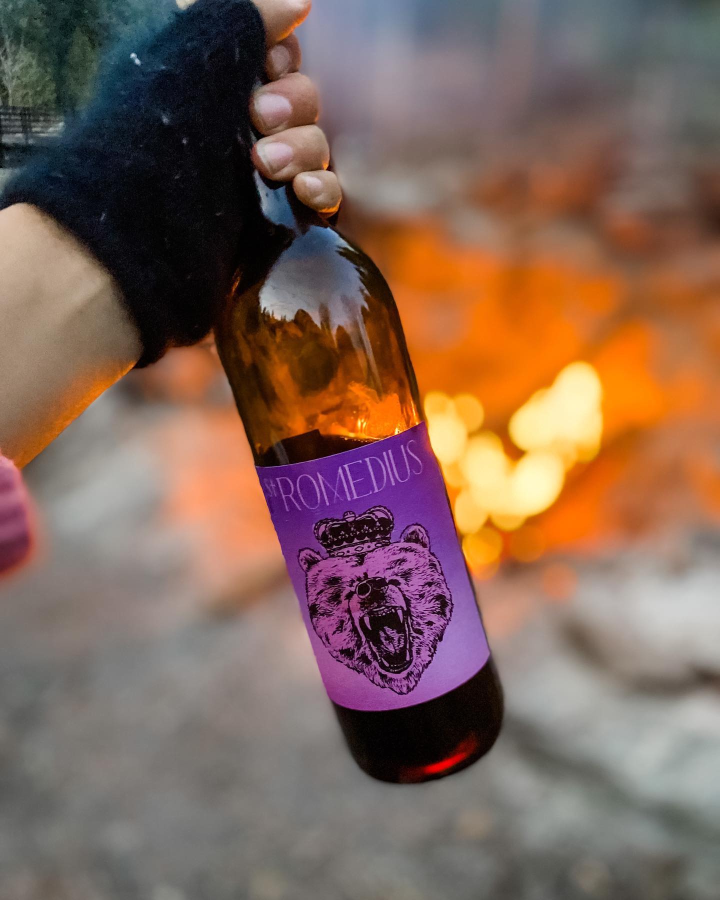 St. Romedius Teroldego wine bottle held by a person wearing gloves, with a campfire burning in the background outdoors.