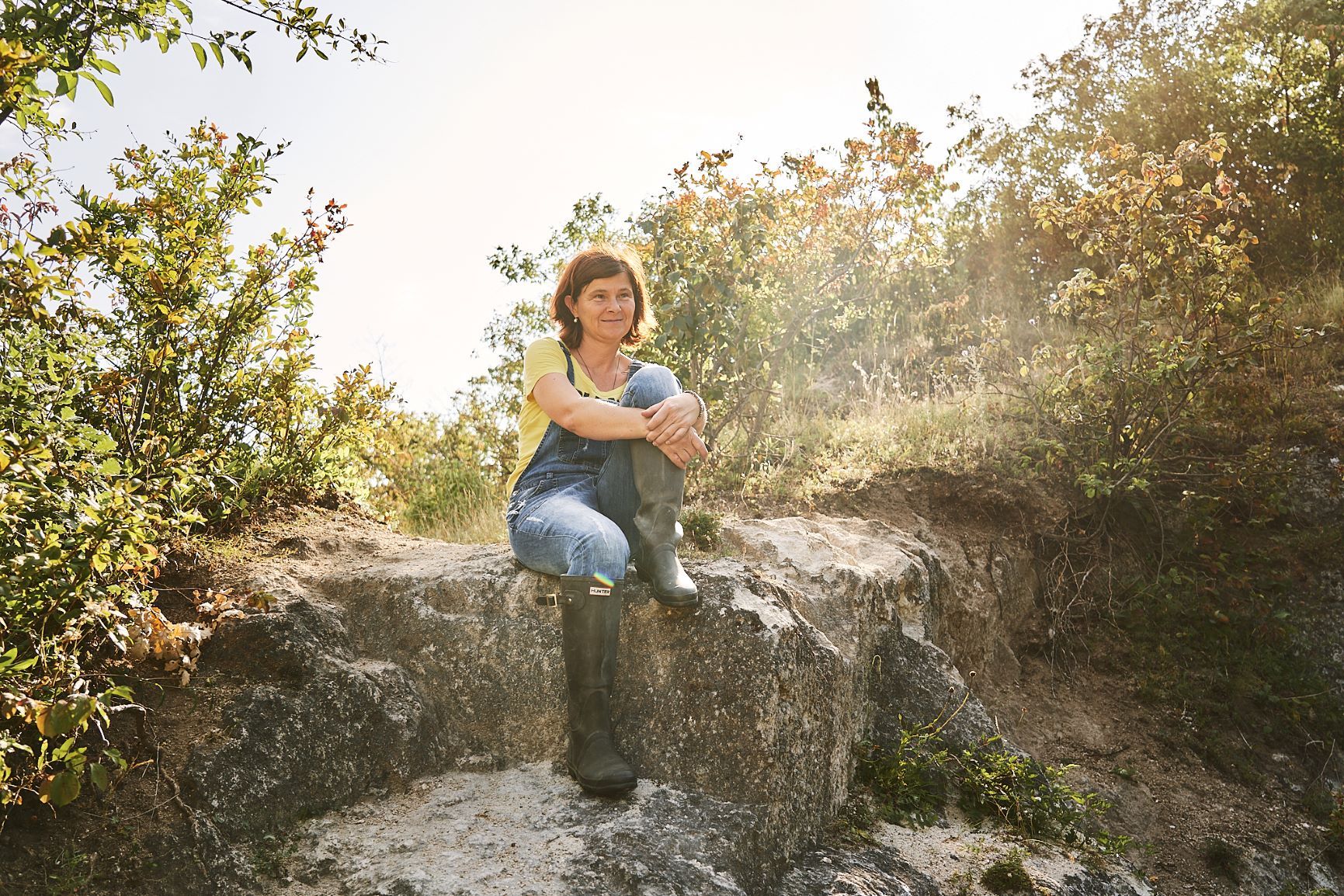 Birgit Braunstein in overalls and boots sits smiling on a rocky ledge, surrounded by foliage and sunlight.