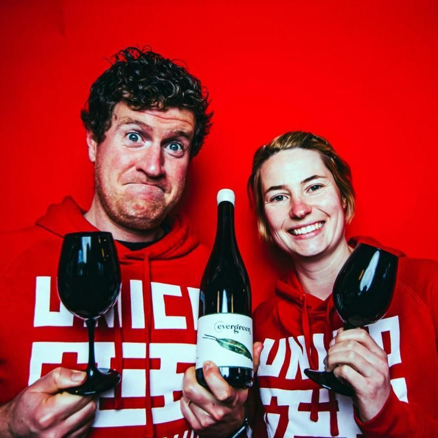Two Unico Zelo winemakers holding wine glasses and a bottle against a red background.
