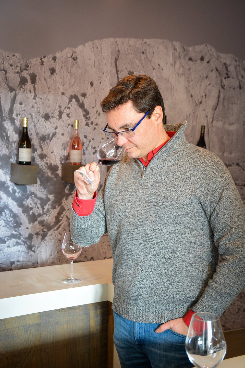 Man sniffing wine in a winery. He wears glasses, a sweater, and jeans. Bottles on shelf in background.