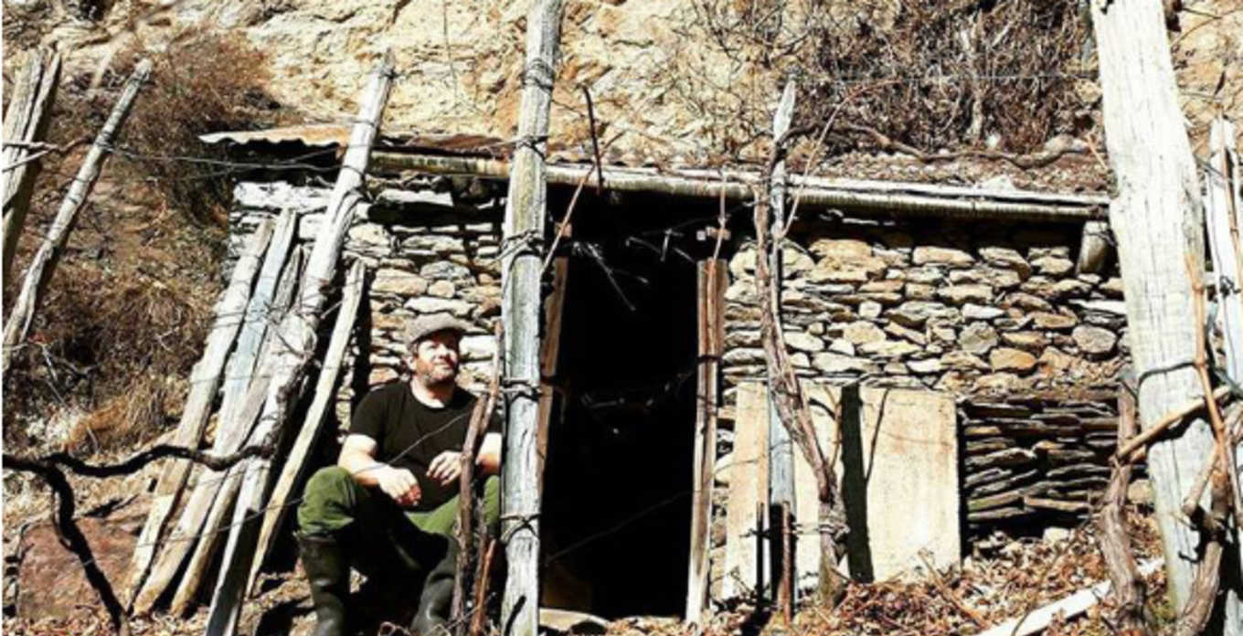 Man sits outside a stone structure built into a hillside, with dry vines and wood framed in the entrance.