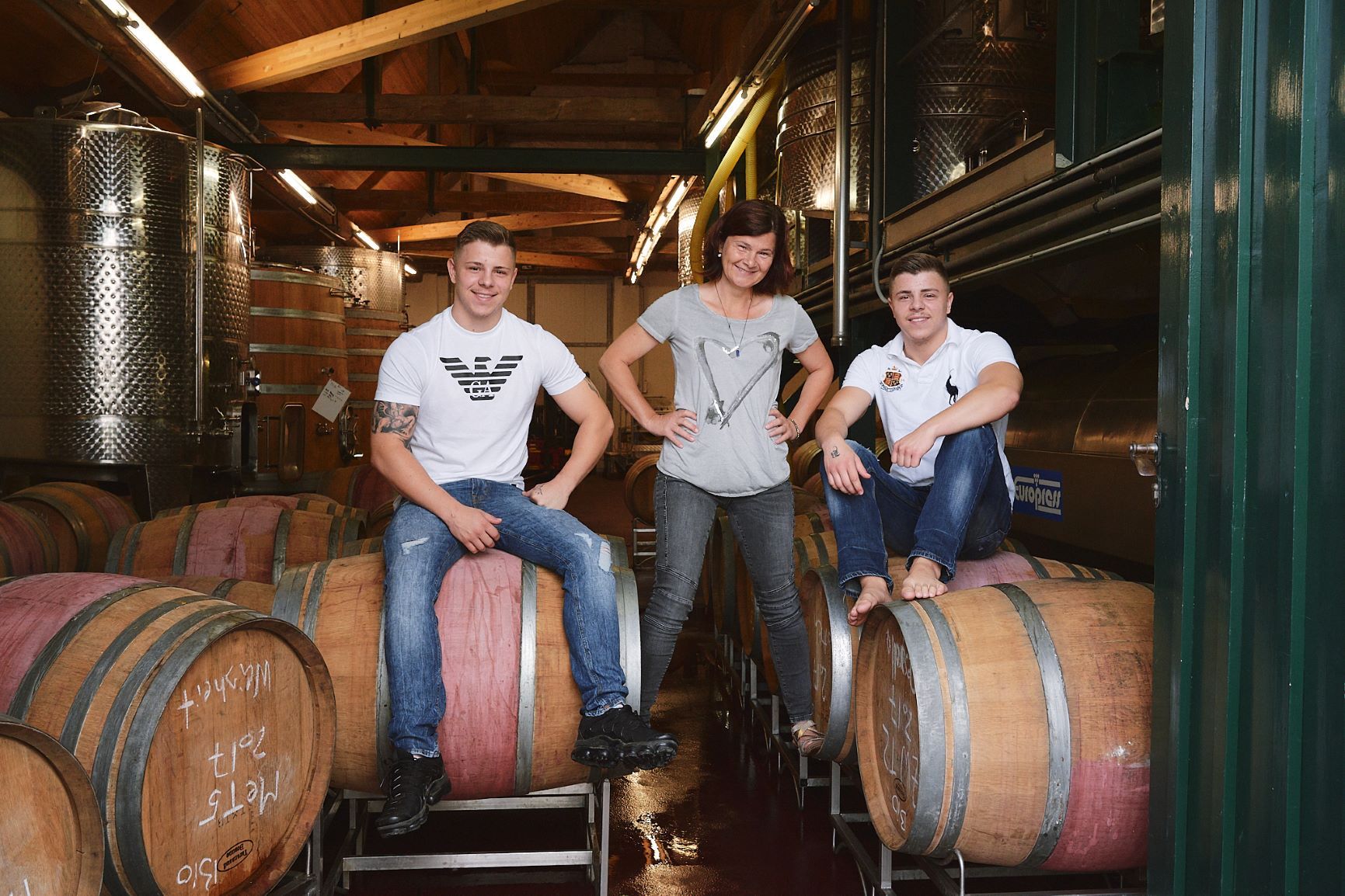 Three people pose in Birgit Braunstein wine cellar. Woman stands, while the two men sit on barrels.