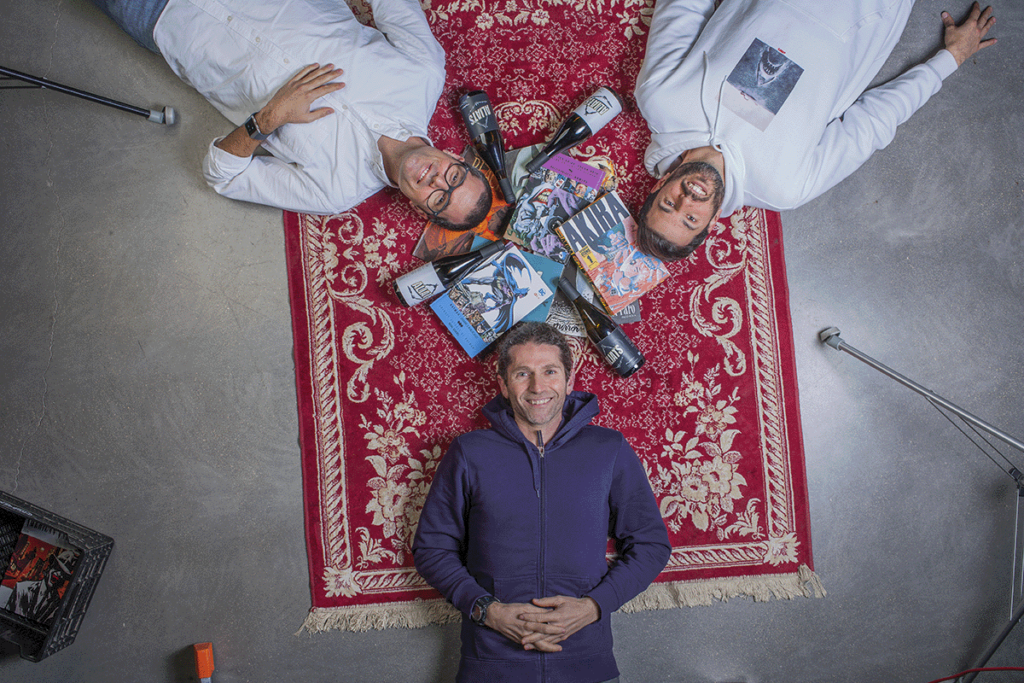 Three men lying on a red rug, at Bodegas Enguera, with four wine bottles and books at the center.
