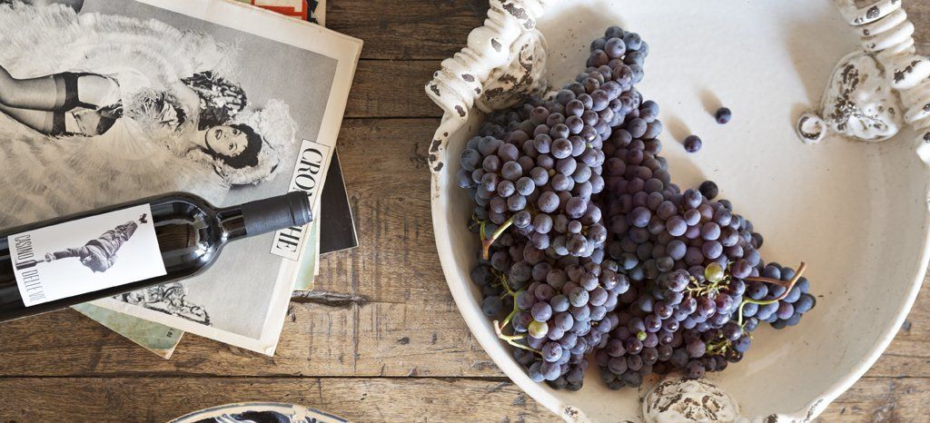 Grapes in a decorative white dish, wine bottle, and vintage photos on a wooden table.