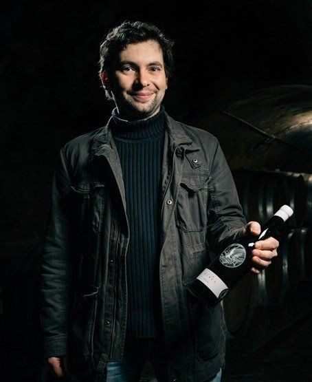A winemaker from Zillinger stands in a cellar holding a bottle of wine, with oak barrels in the background.
