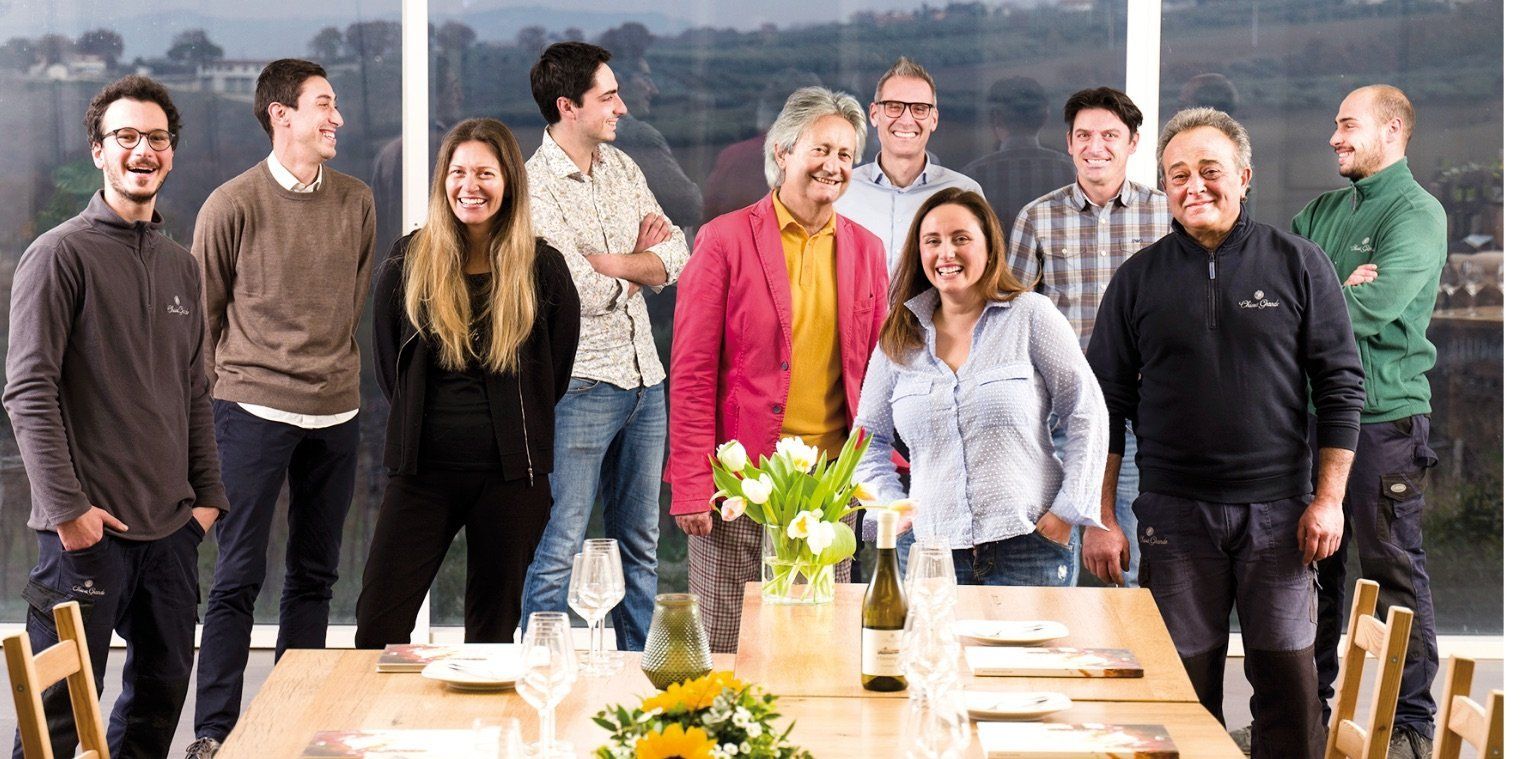 Group of people smiling around a table at Chiusa Grande, with a vineyard visible in the background.