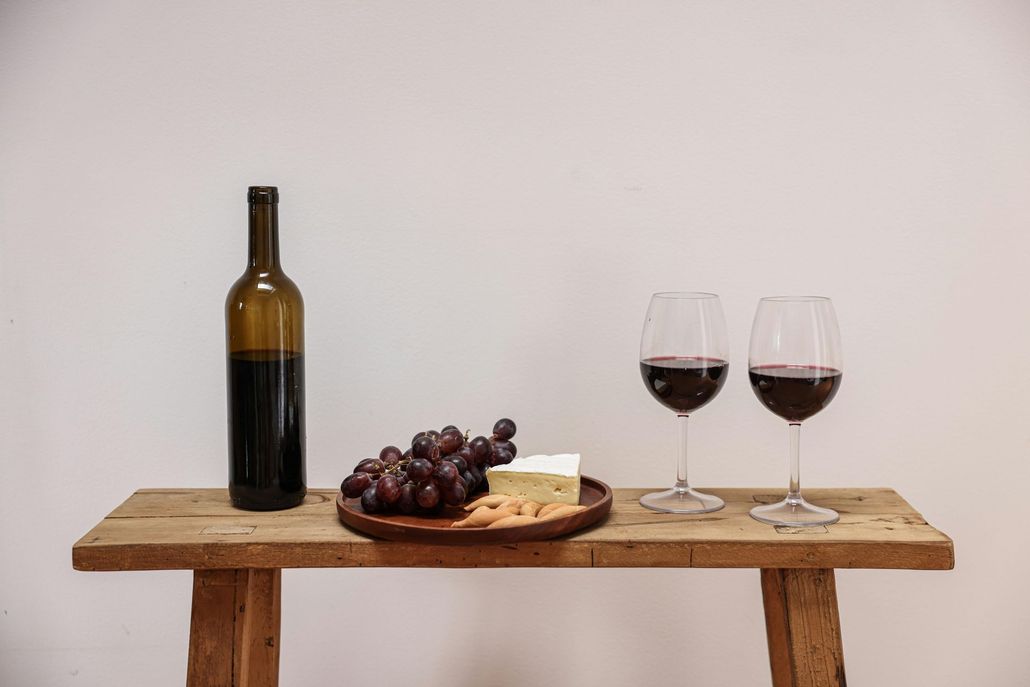 Wine bottle, glasses, grapes, and cheese on a wooden table against a white wall.