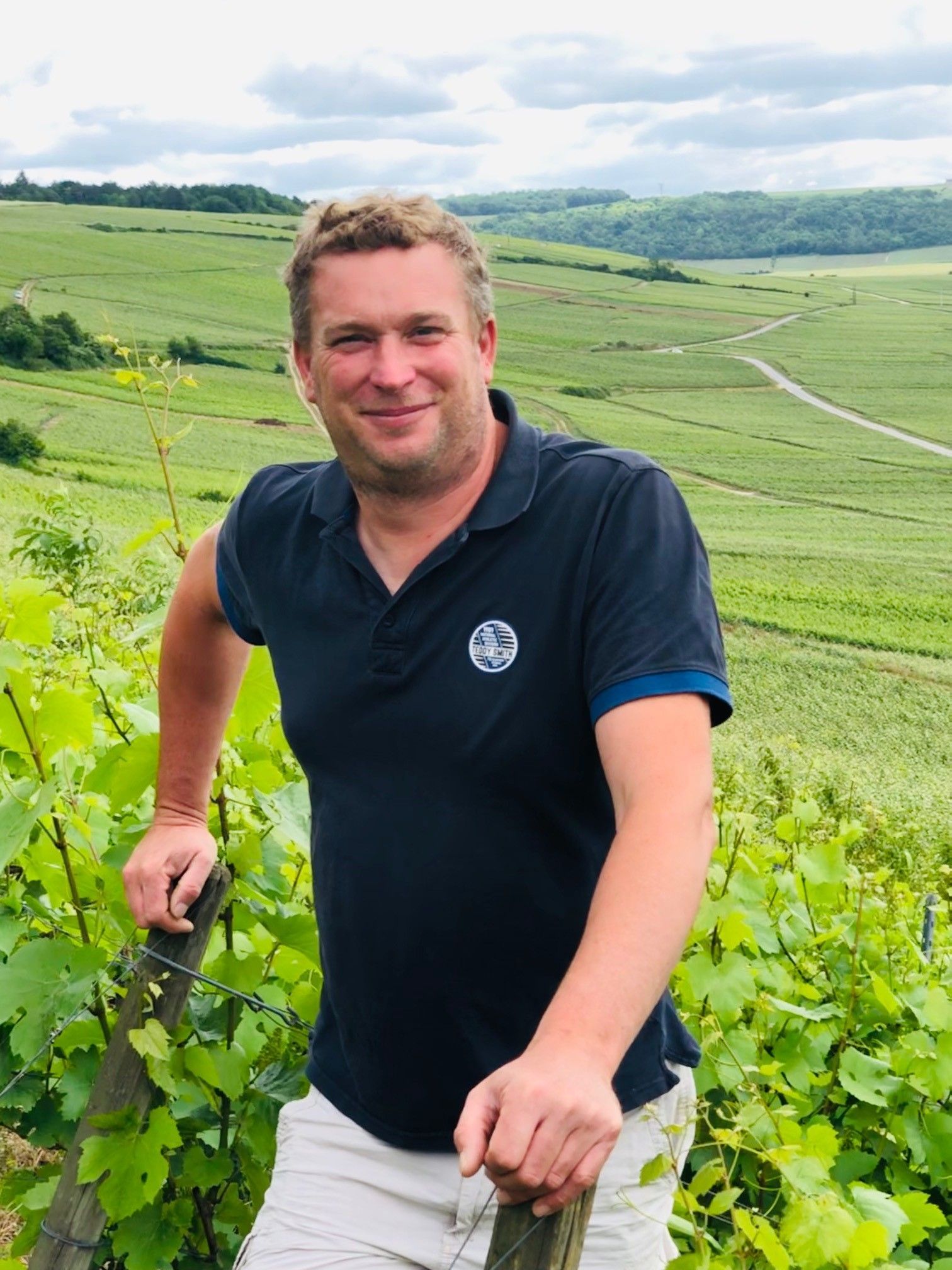 Man smiling in Champagne Olivier Marteaux vineyard, rolling hills in the background.