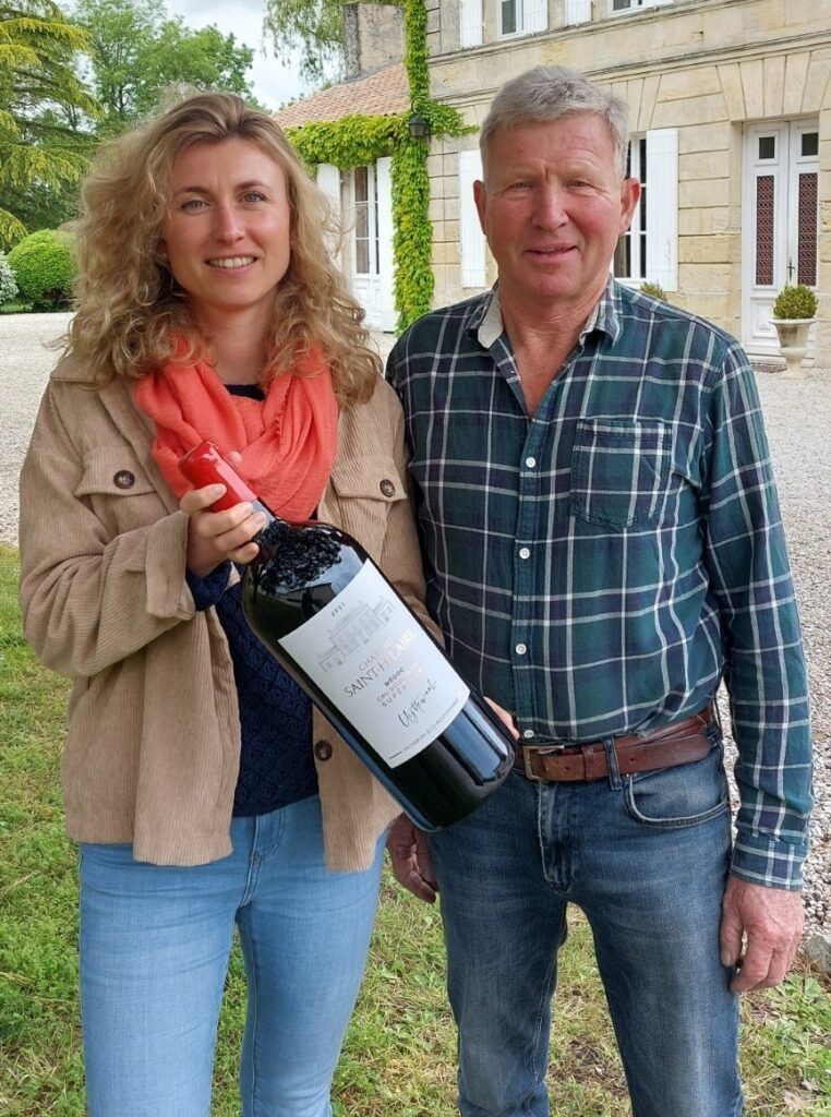 Woman and man holding a large wine bottle of Château Saint Hilaire wine near a stone building.