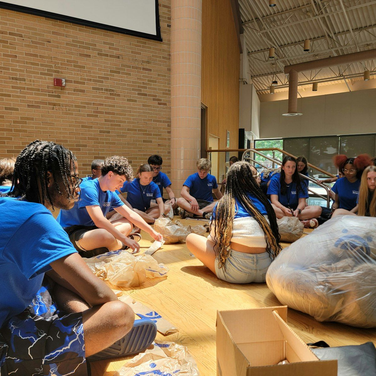 A group of people wearing blue shirts sit on a wooden floor in a large room, sorting items from plastic bags into boxes.