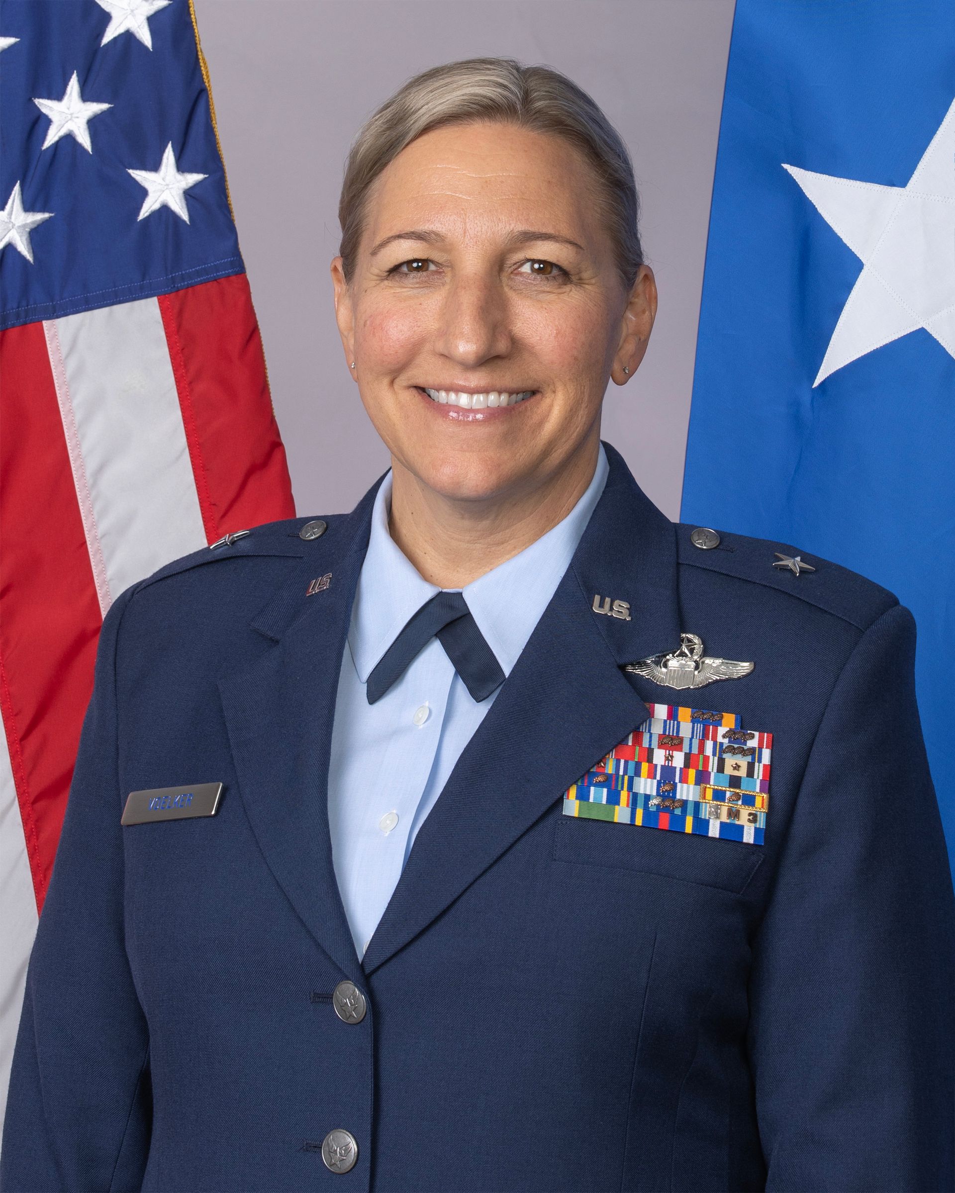 A US Air Force officer in uniform stands before the American flag and a blue general officer flag.