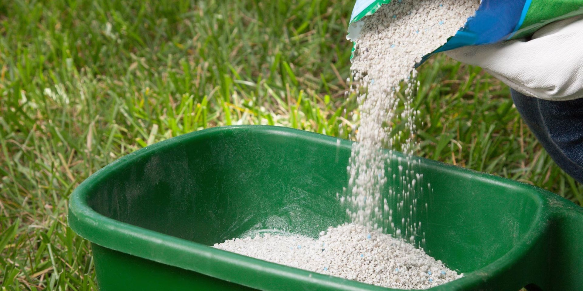 A person is pouring fertilizer into a green wheelbarrow.