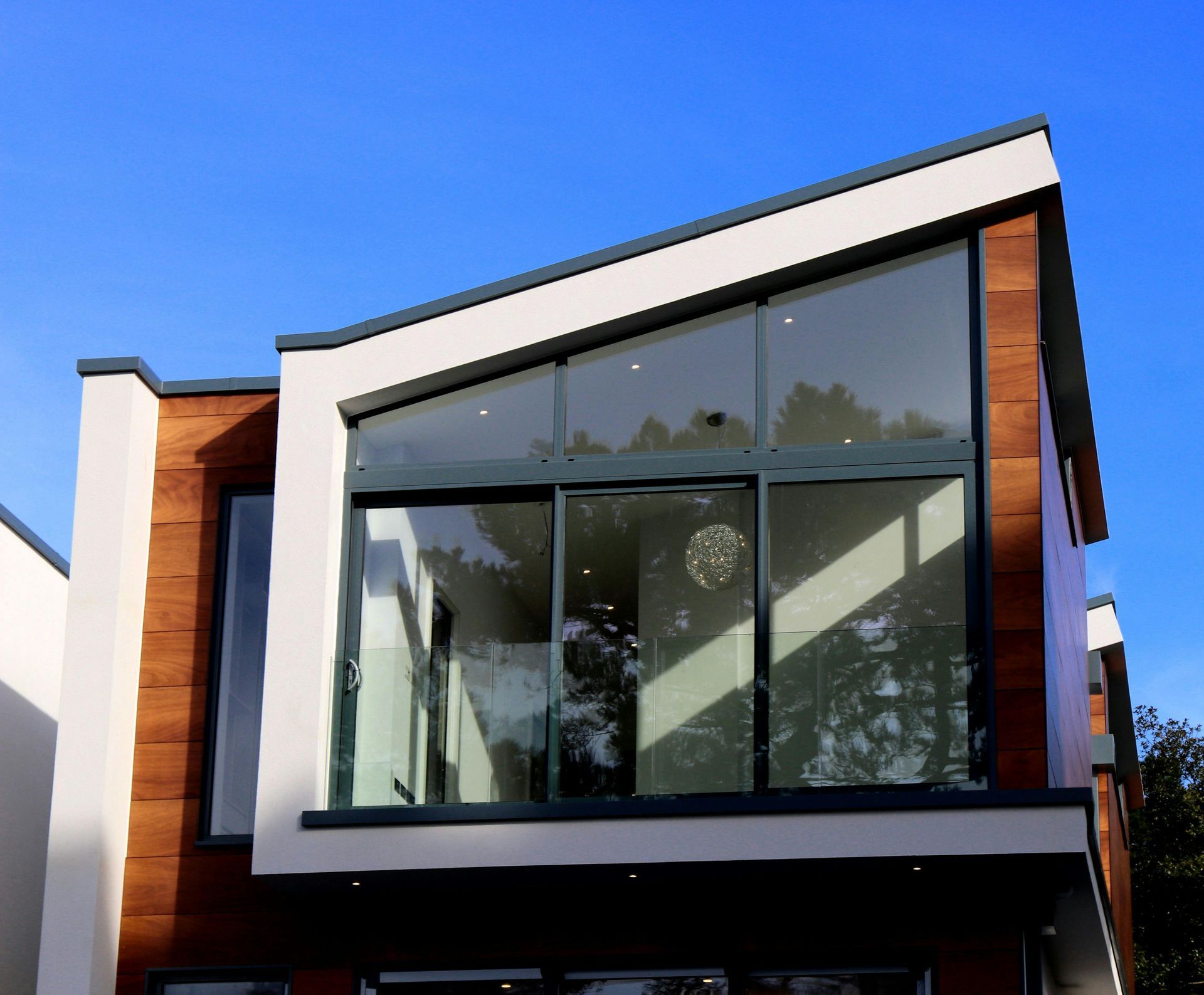 Modern house exterior with white stucco, wood paneling, and a large angled glass window against a clear blue sky.