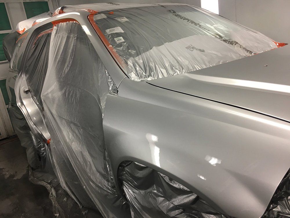 A silver car is covered in plastic while being painted in a garage.