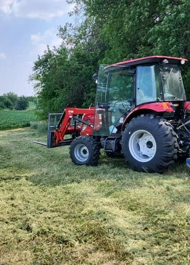 A red tractor with a forklift attached to it is cutting grass in a field.