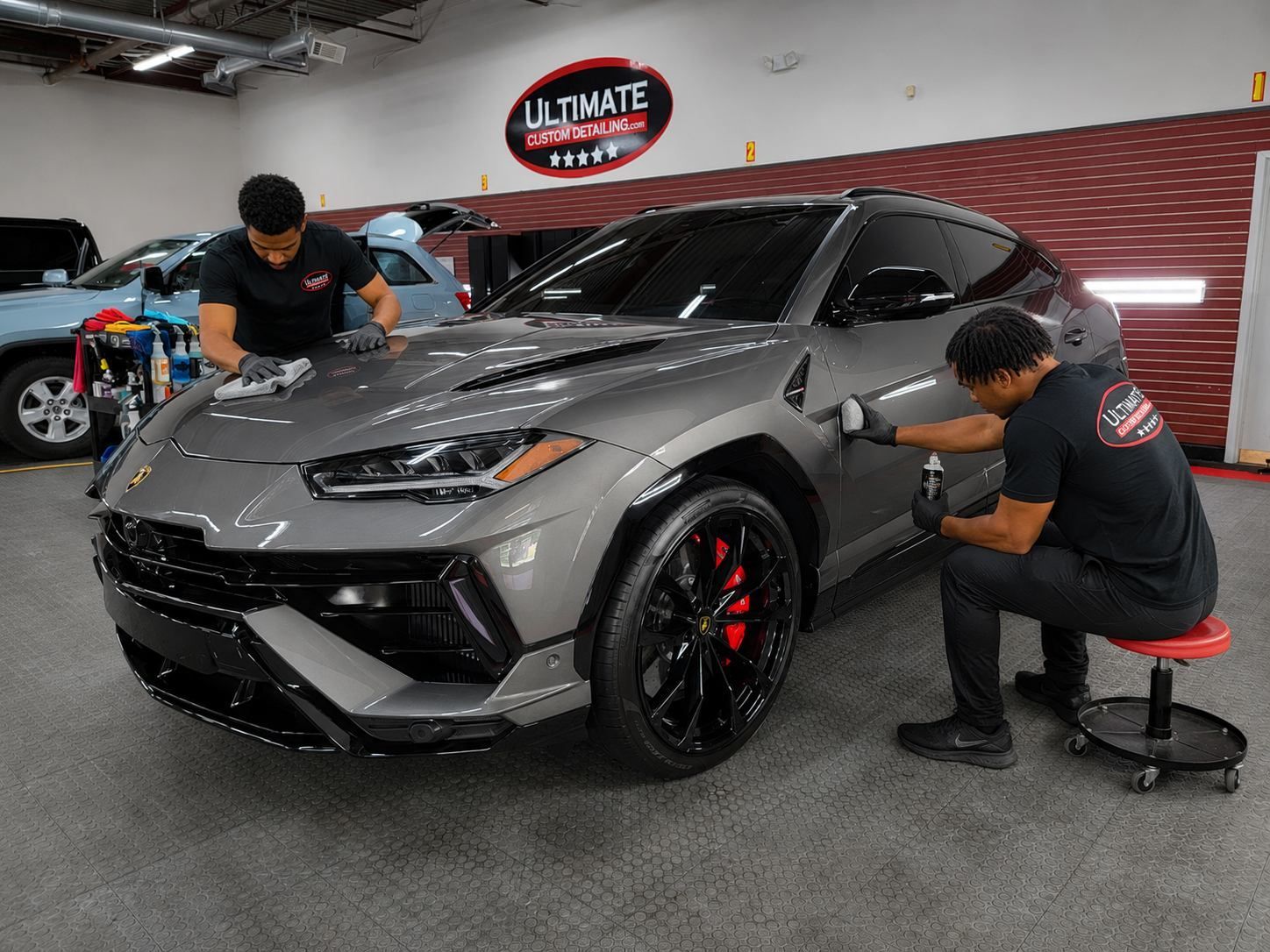 Two detailers polishing a gray sports car inside an auto shop