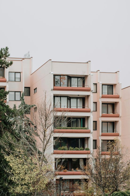 Un immeuble d'appartements rose pâle avec des balcons étagés et de grandes fenêtres, entouré d'arbres par temps nuageux.