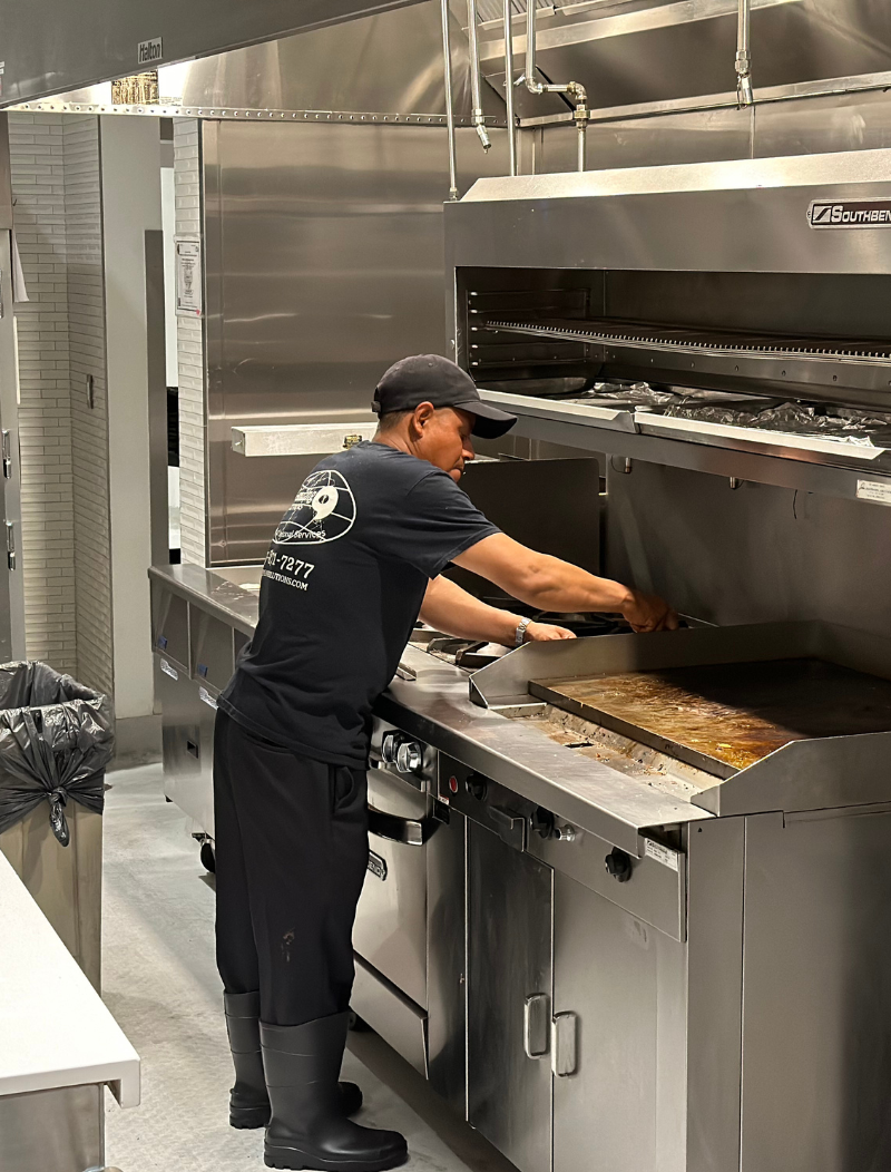 A man is standing in a kitchen scraping and cleaning a grill in preparation for sanitization.