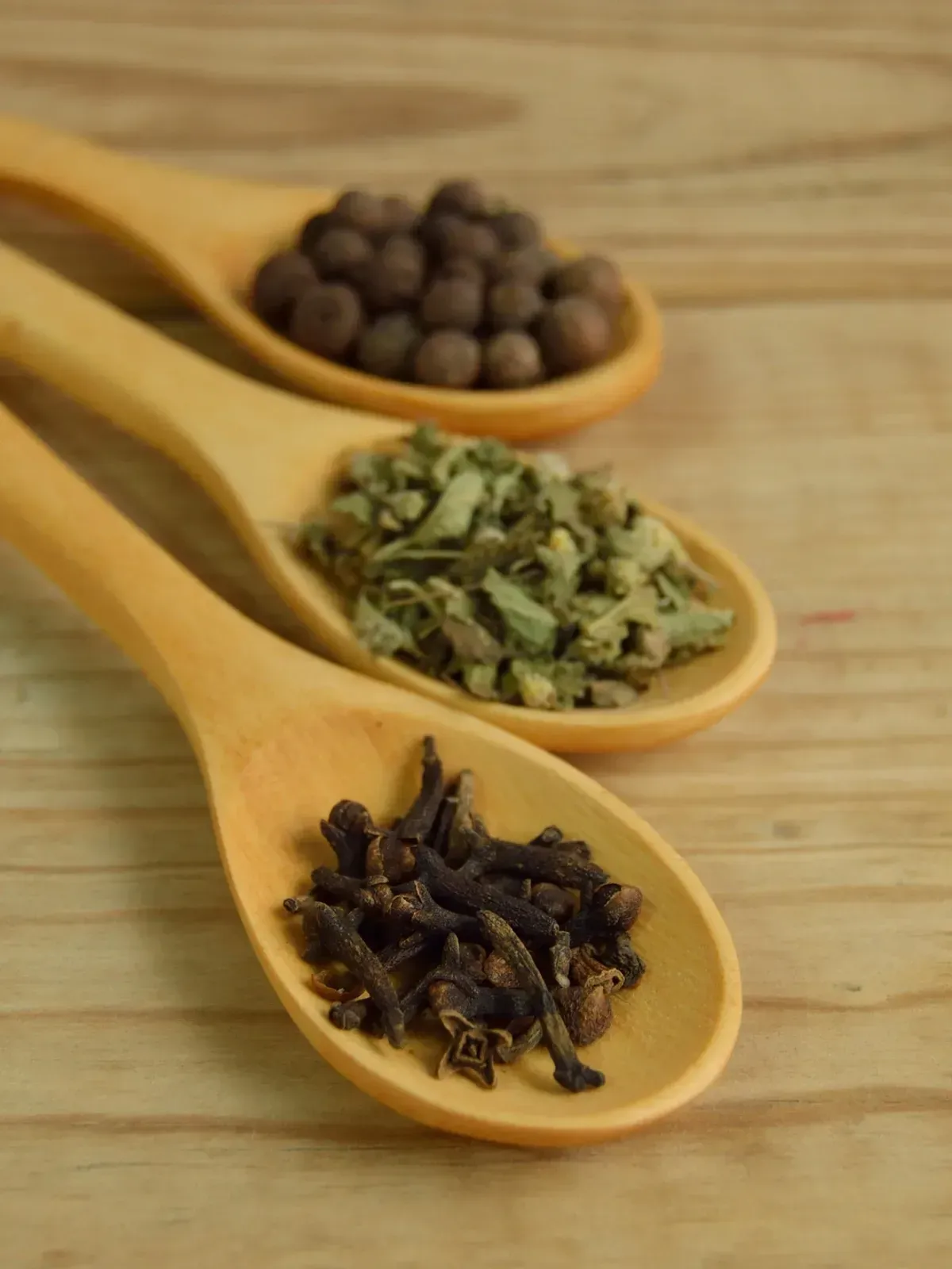 Three wooden spoons holding dried herbs and spices on a wooden surface