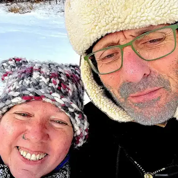 Two people smiling outdoors in winter hats, close together in snowy surroundings.
