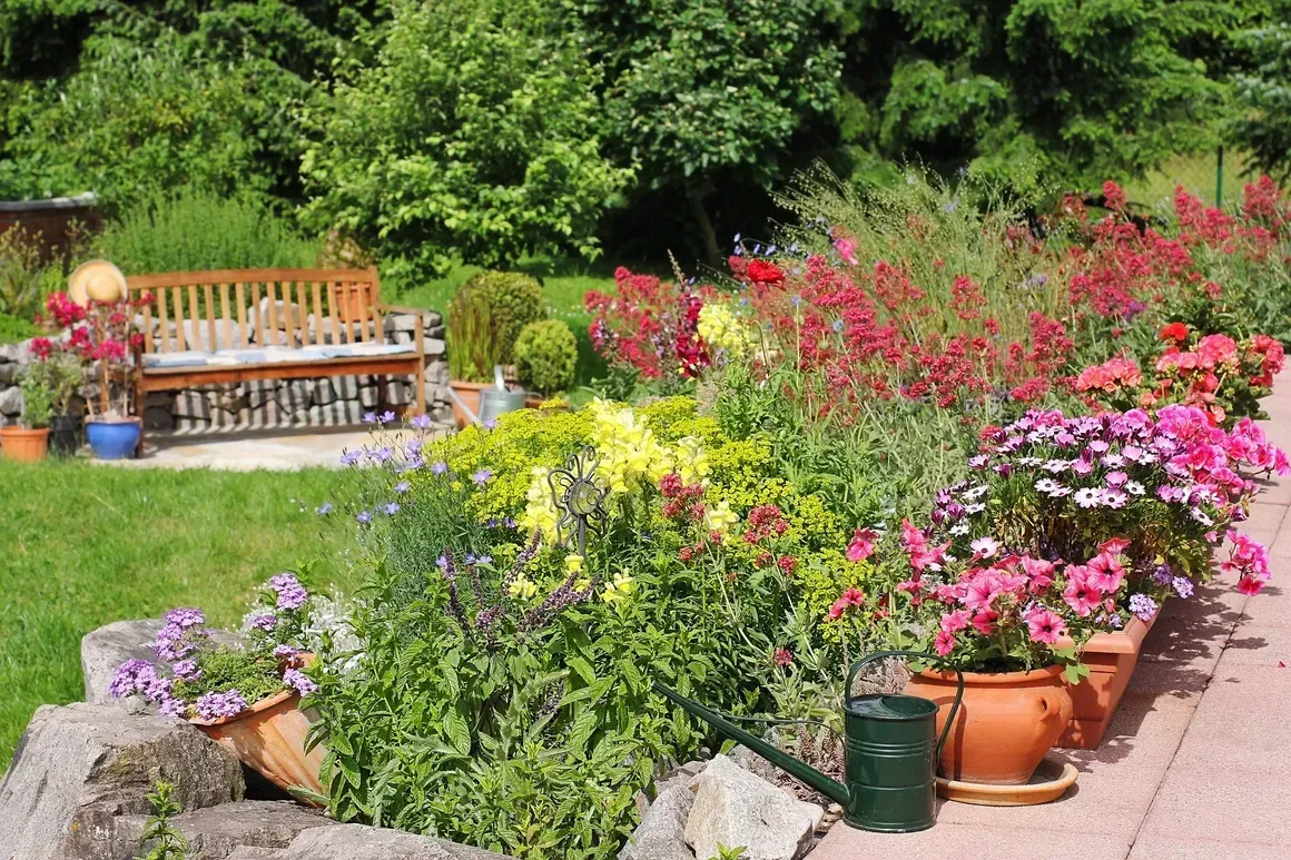 Colorful flowers in terracotta pots line a garden path beside a wooden bench and lush green lawn.