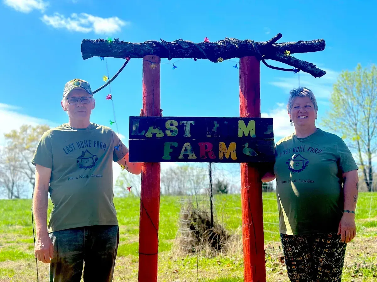 Two people standing beside a Last Farm sign in a sunny field under a blue sky