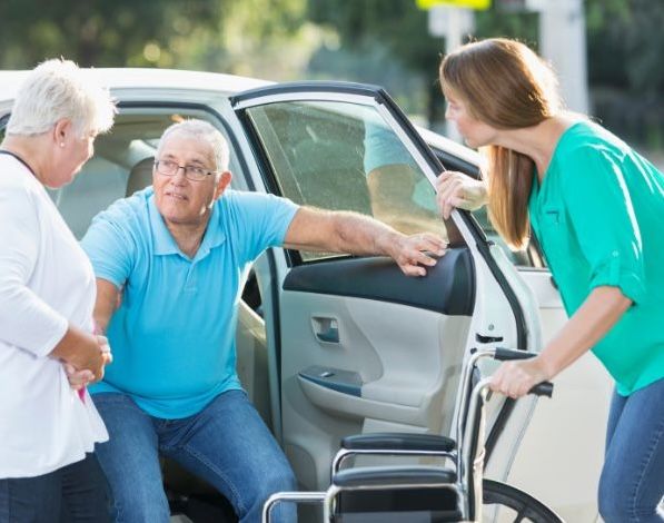Woman and aide helping man get out of car into a wheelchair