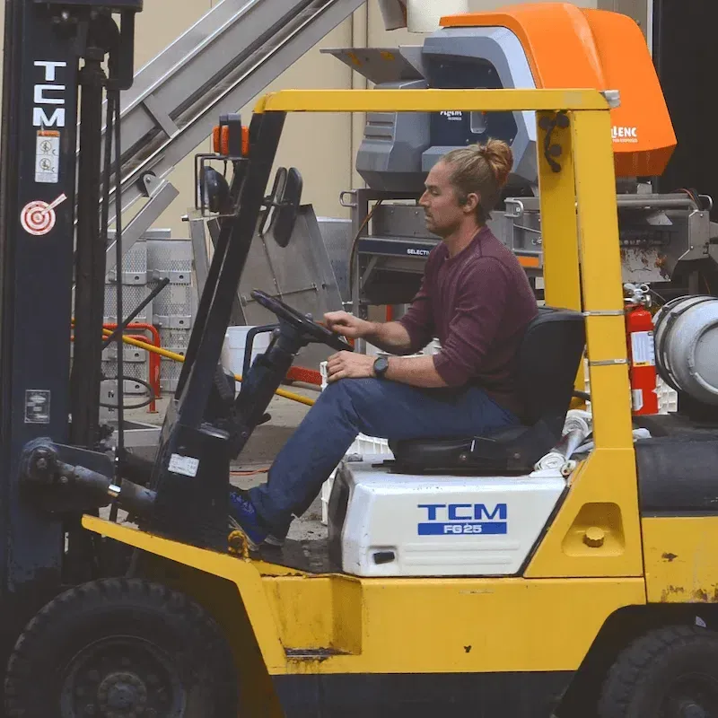 A man driving a yellow forklift at a winery in California, Napa, with equipment and barrels in the background.