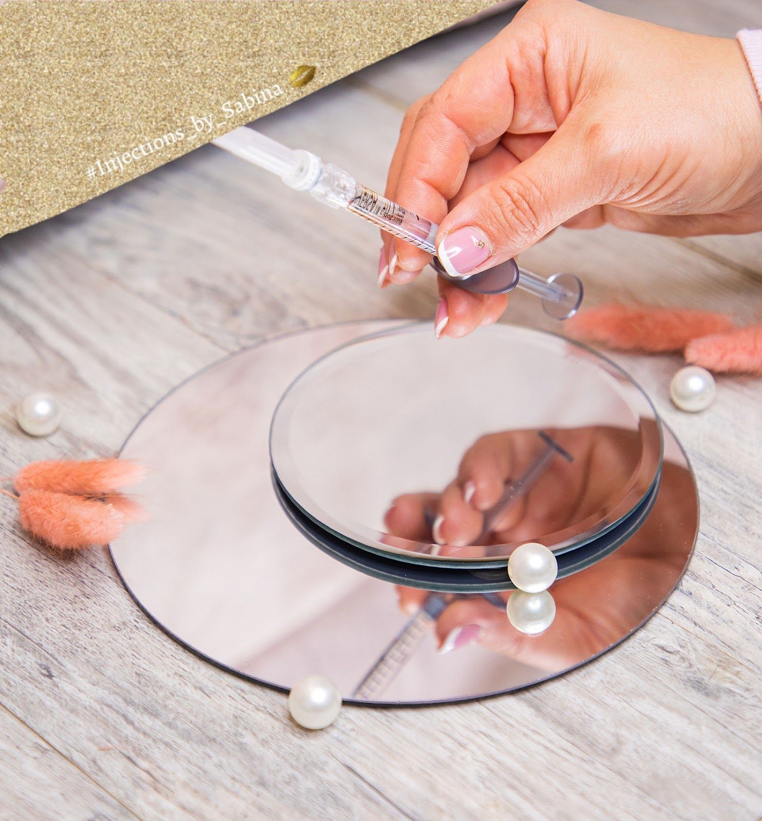 Hand holding syringe over a mirrored surface with pearls and pink decor on a wooden surface.