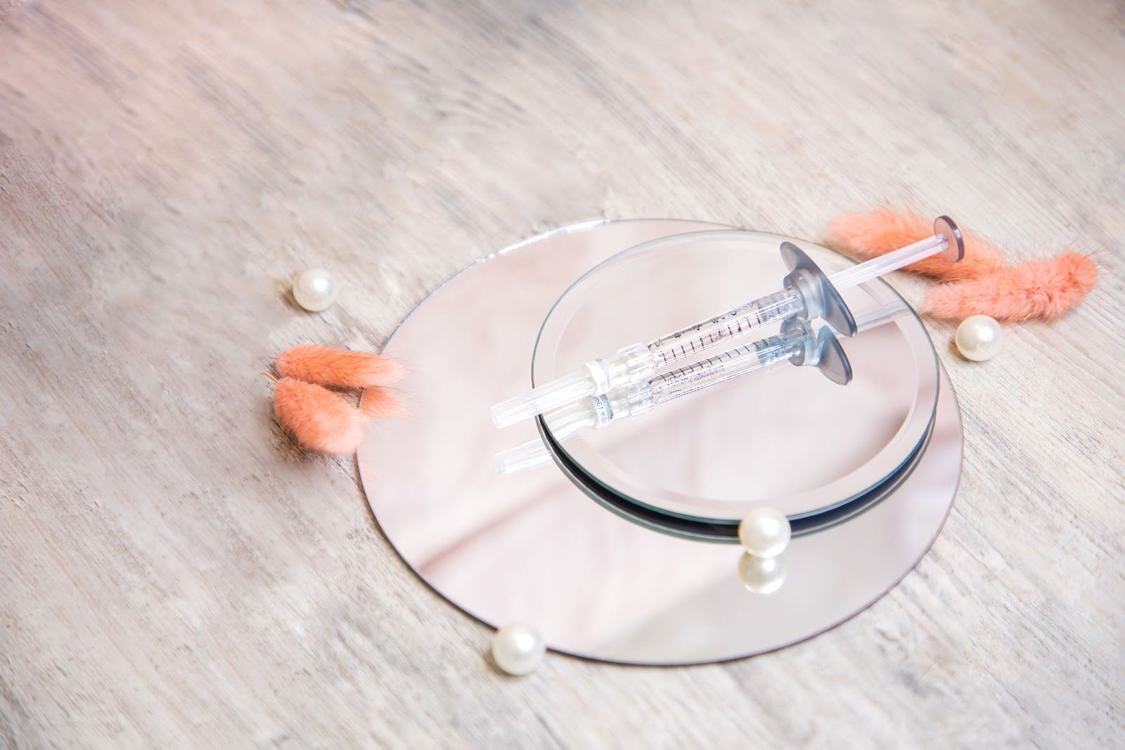 Syringe on reflective circles, pearls, and pink accents on a light wood surface.