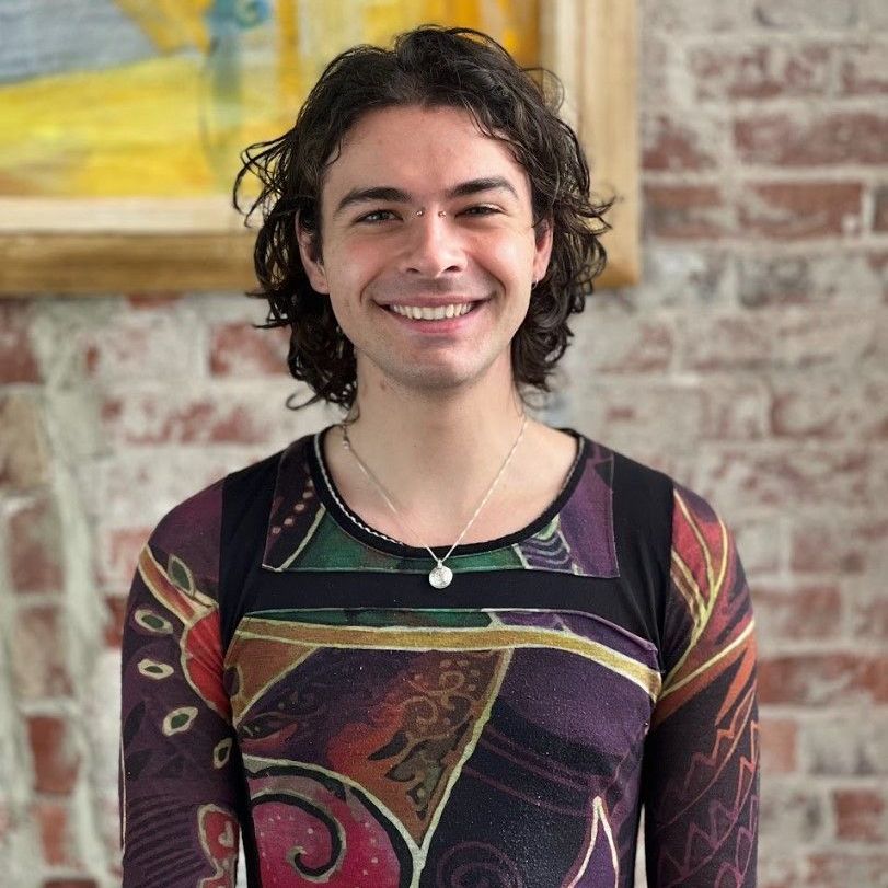 Man with curly dark hair smiles at the camera, wearing a colorful long-sleeved shirt, against a brick wall.