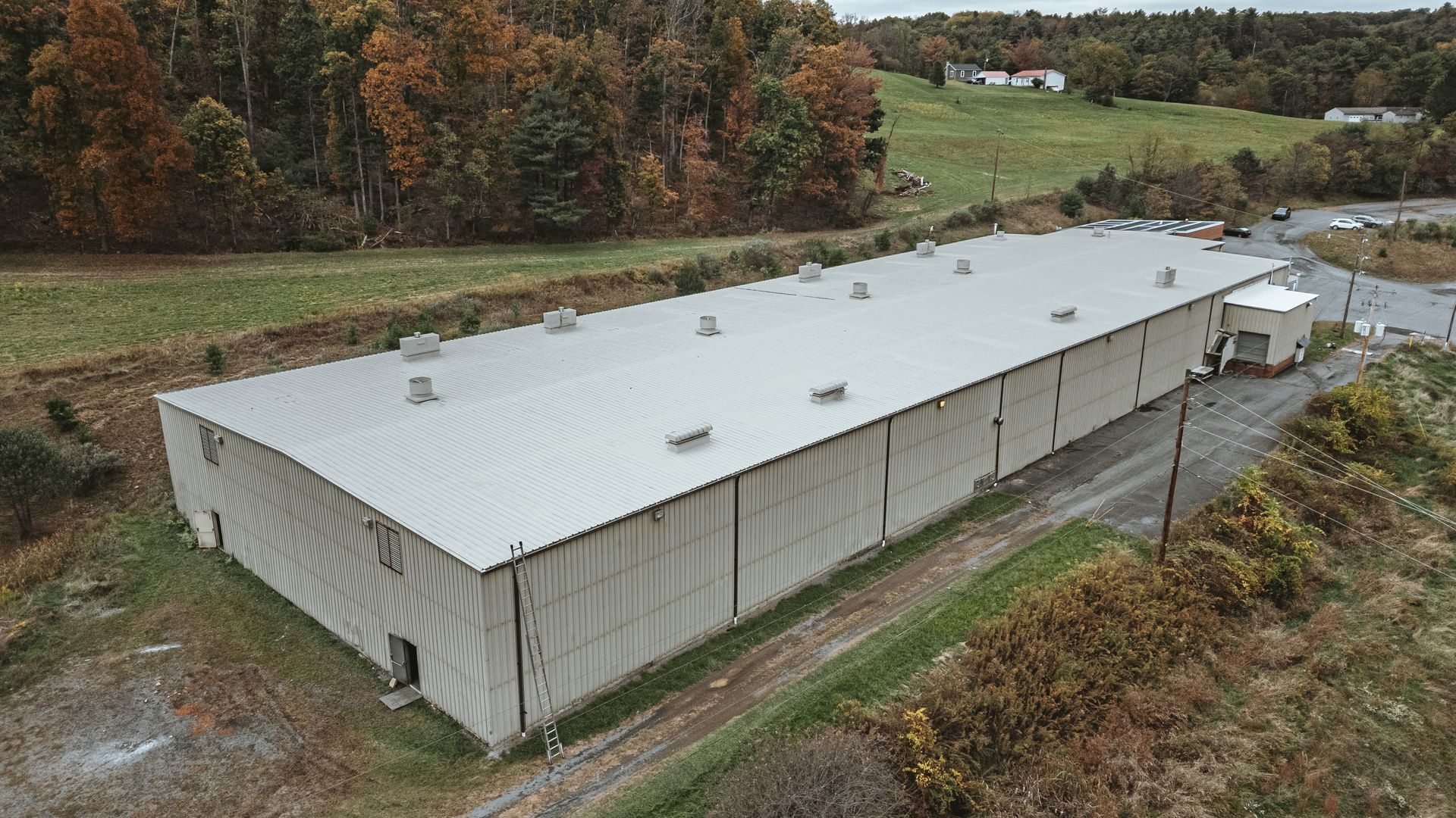 Large, gray industrial building with a corrugated metal roof, surrounded by trees and a grassy field.