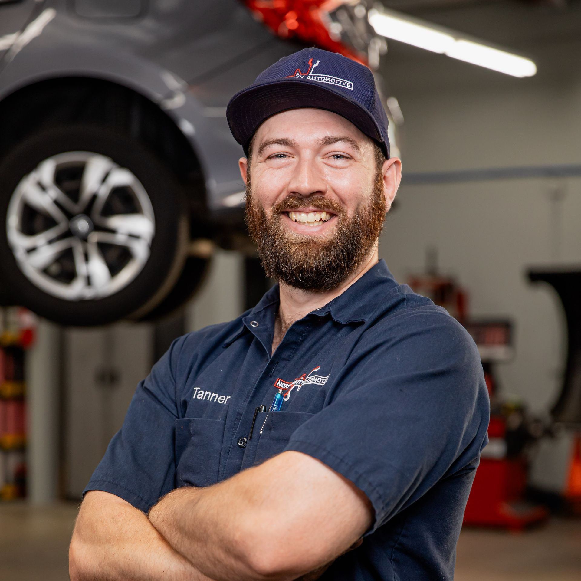 A man with a beard is standing in front of a car in a garage.