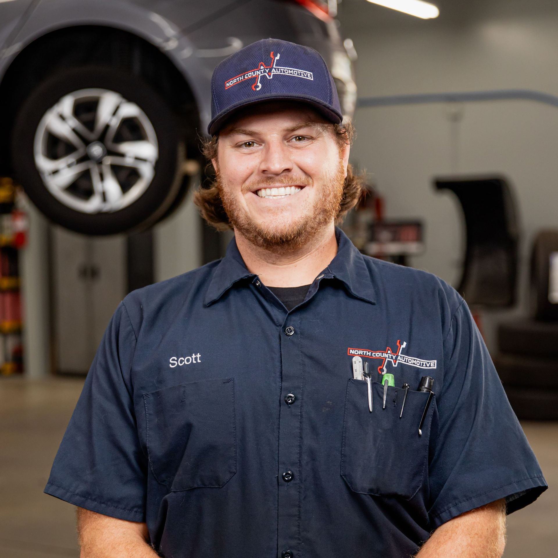 A man in a blue shirt and hat is standing in front of a car in a garage.