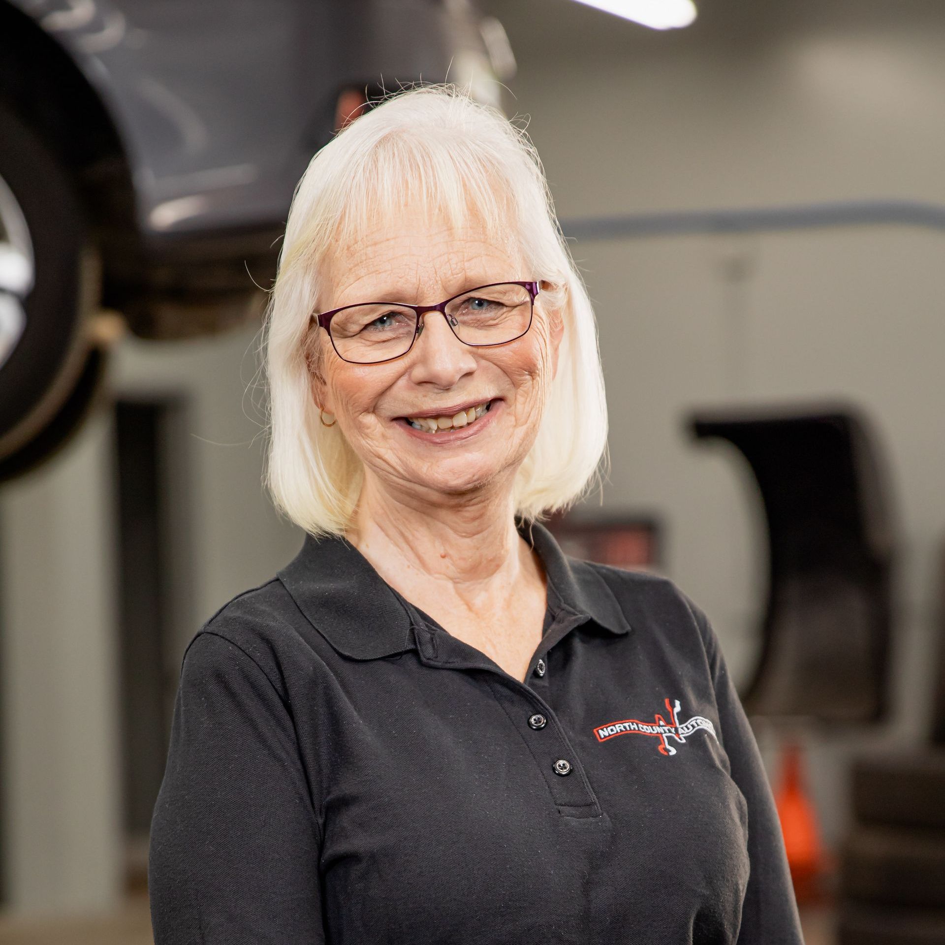 A woman wearing glasses and a black shirt is smiling in front of a car.