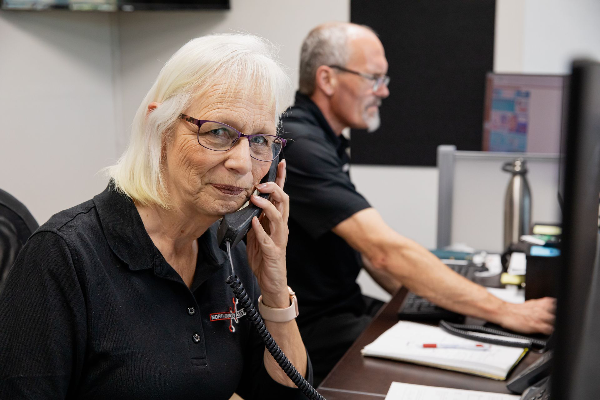 A woman is talking on a phone while a man is using a computer.