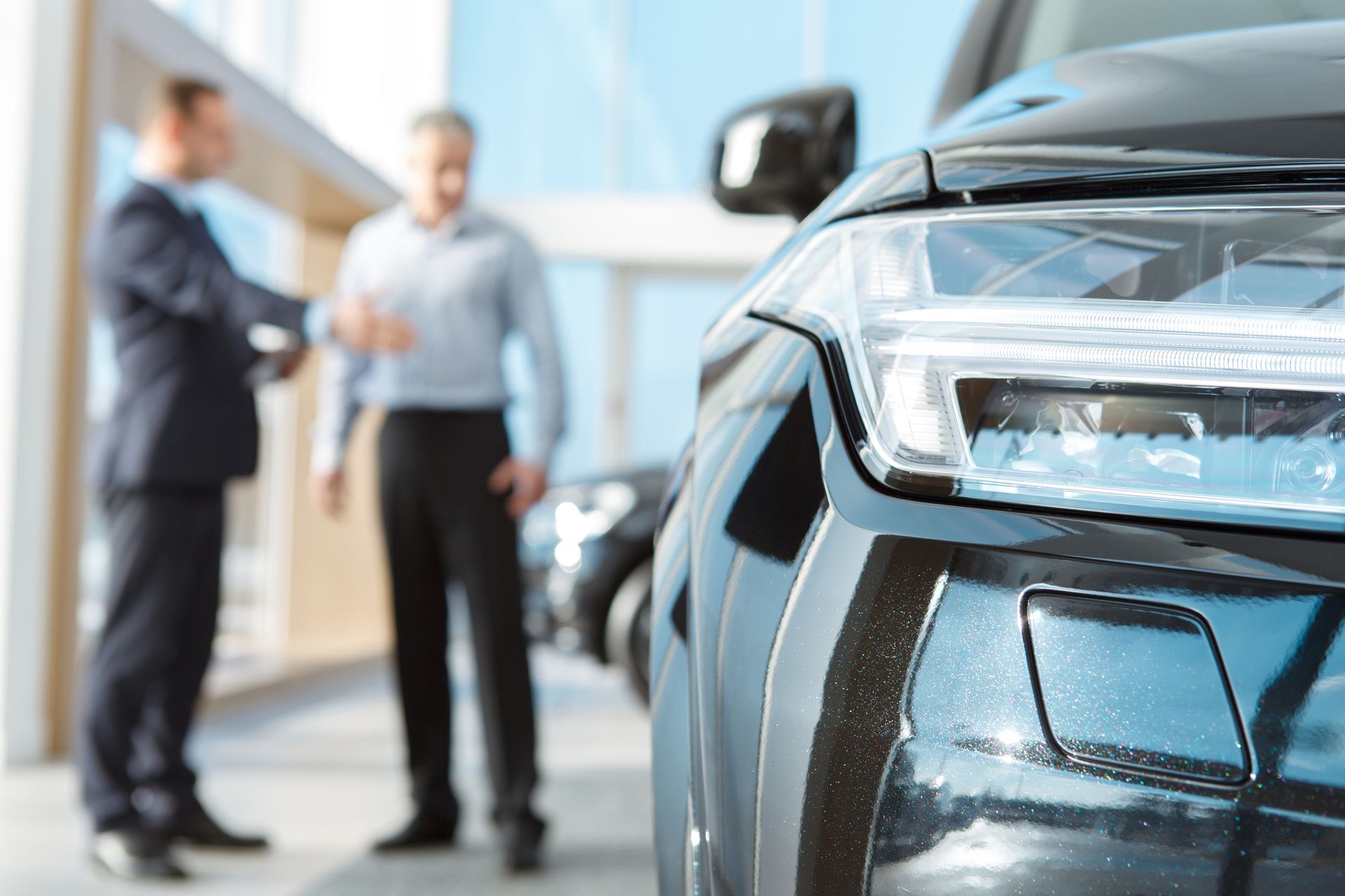 Close-up of a shiny black car with two blurred people talking beside it outside a dealership