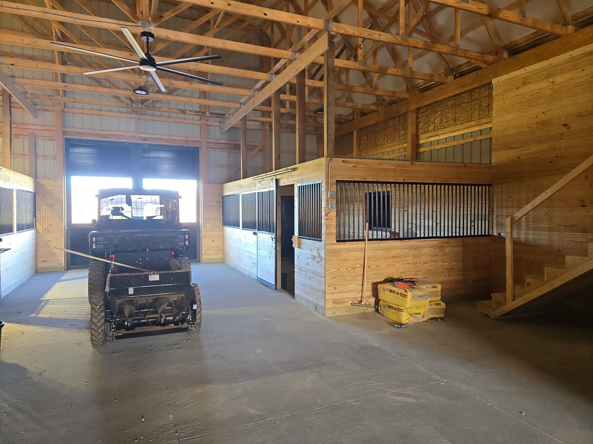 An indoor barn with a UTV, several horse stalls, a loft area, and wooden walls under a high exposed-beam ceiling.
