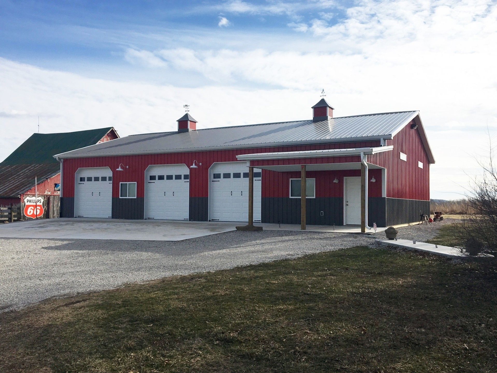 Red and gray barn with three garage doors, a carport, and a metal roof against a blue sky.
