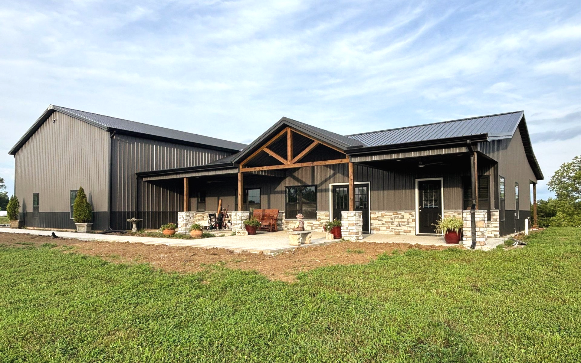 Brown barn-style building with a porch and stone accents, set in a grassy field under a blue sky.
