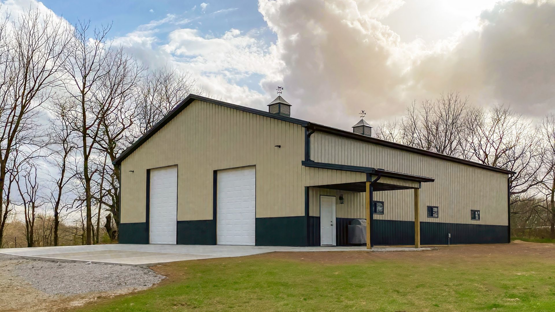 Tan metal building with white doors and a small porch on a grassy lot under a cloudy sky