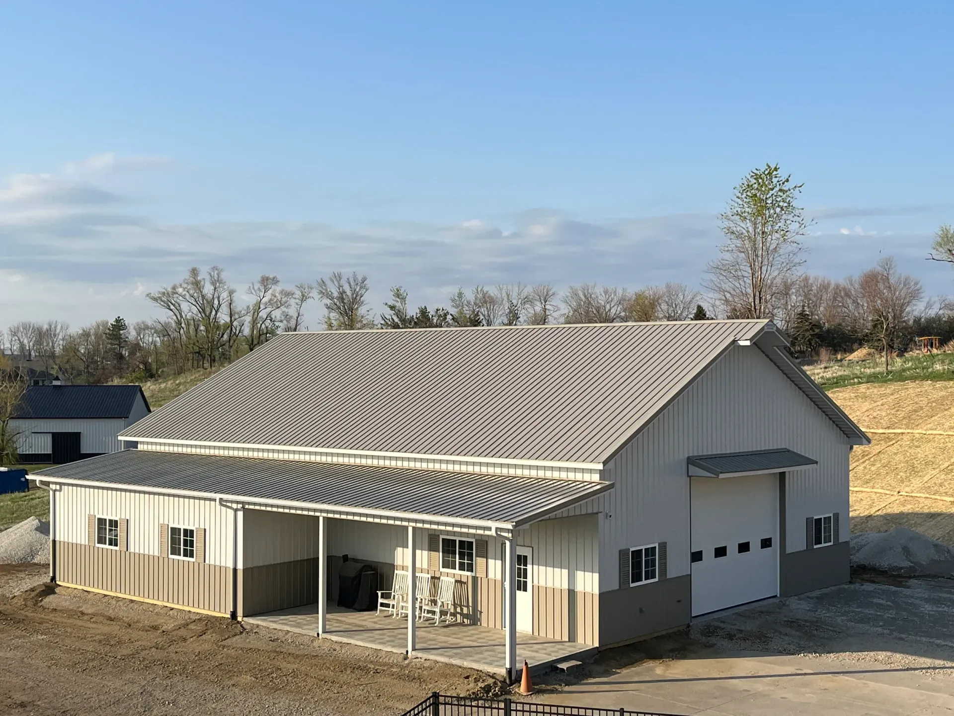 White and tan metal building with a covered porch and garage door, set in a rural landscape under a blue sky.